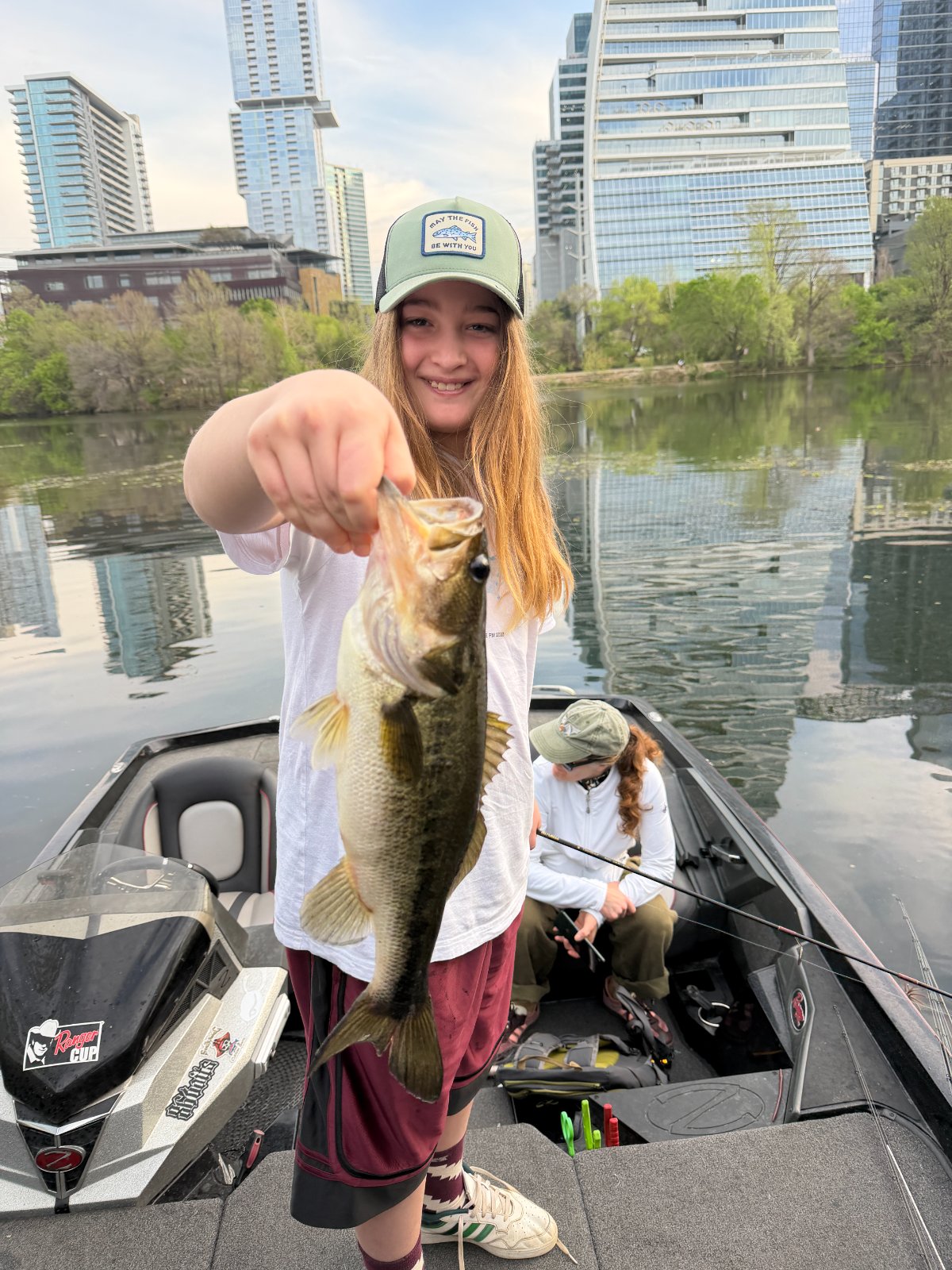 A young angler holding out a largemouth bass with the downtown Austin skyline behind